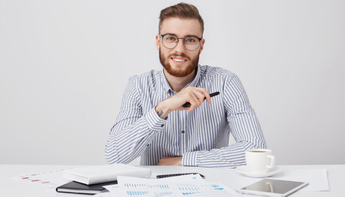 Professional male worker with thick beard and trendy hairstyle, wears round glasses and formal shirt, sits at work plae, drinks coffee, works with documents. Hipster young guy being busy working