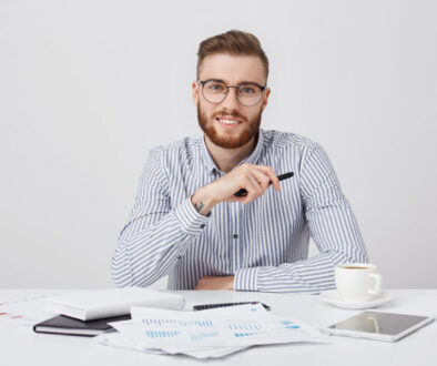 Professional male worker with thick beard and trendy hairstyle, wears round glasses and formal shirt, sits at work plae, drinks coffee, works with documents. Hipster young guy being busy working